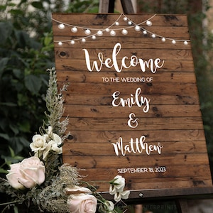 May include: Rustic wooden wedding welcome sign with string lights, featuring the text "Welcome to the wedding of Emily & Matthew, September 18, 2023".