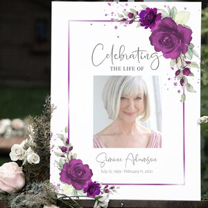 May include: A white memorial sign with a purple floral border and a photo of a woman. The text reads "Celebrating the Life of Simone Adamson July 15, 1959 - February 11, 2021".