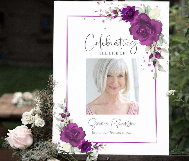 May include: A white memorial sign with a purple floral border and a photo of a woman. The text reads "Celebrating the Life of Simone Adamson July 15, 1959 - February 11, 2021".