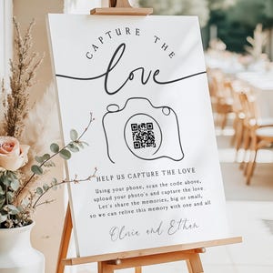 May include: A white rectangular wedding sign on a wooden easel featuring the black text CAPTURE THE LOVE, a line-art camera illustration with a central QR code, and the names Olivia and Ethan, along with instructions for guests to upload photos.