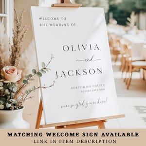 May include: A white wedding welcome sign on a wooden easel. The sign reads "Welcome to the Wedding of Olivia and Jackson" with the wedding date and location. The bottom of the sign says "we're so glad you're here!"