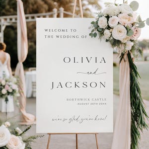 Op de afbeelding: Een wit welkomstbord voor een bruiloft op een gouden ezel, versierd met bloemstukken. Op het bord staat "WELCOME TO THE WEDDING OF OLIVIA and JACKSON" met de trouwlocatie en -datum. Het bord zegt ook "we're so glad you're here!"