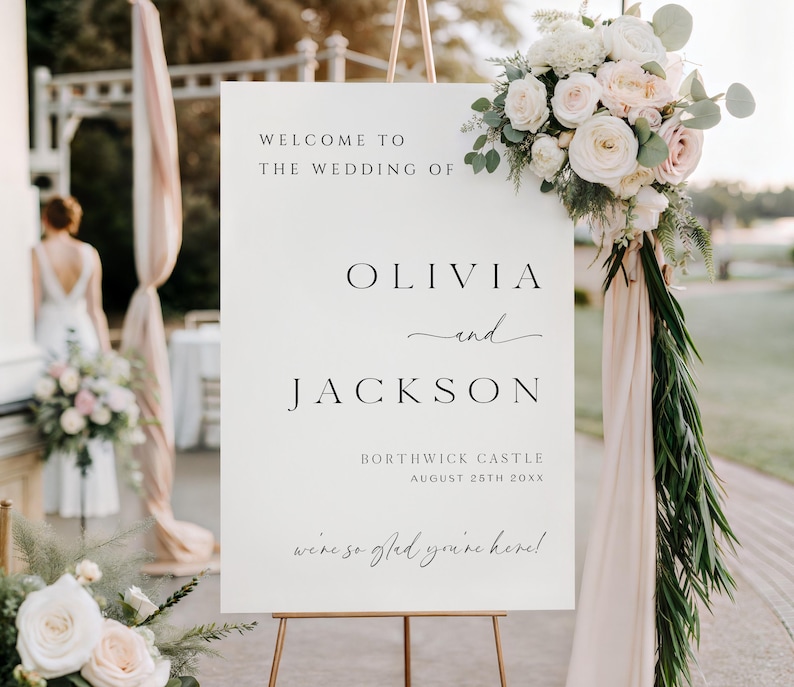 Op de afbeelding: Een wit welkomstbord voor een bruiloft op een gouden ezel, versierd met bloemstukken. Op het bord staat "WELCOME TO THE WEDDING OF OLIVIA and JACKSON" met de trouwlocatie en -datum. Het bord zegt ook "we're so glad you're here!"