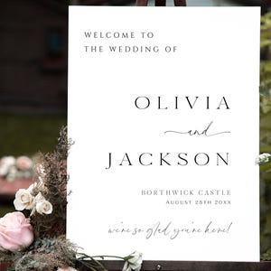 Op de afbeelding: Een wit trouwbord met zwarte tekst. Het bord leest "WELCOME TO THE WEDDING OF OLIVIA and JACKSON" met de locatie en datum hieronder. Het bord is versierd met bloemen.