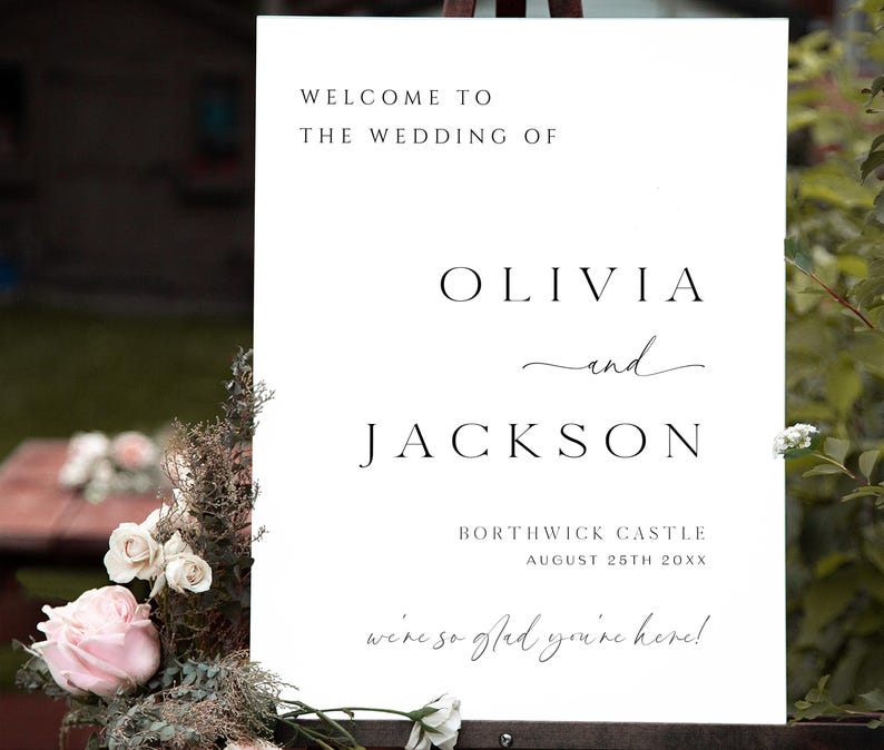 Op de afbeelding: Een wit trouwbord met zwarte tekst. Het bord leest "WELCOME TO THE WEDDING OF OLIVIA and JACKSON" met de locatie en datum hieronder. Het bord is versierd met bloemen.