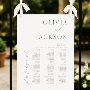 May include: A black metal wedding sign with a white paper insert. The sign features the names "Olivia and Jackson" and a seating chart with guest names. White ribbons adorn the top.