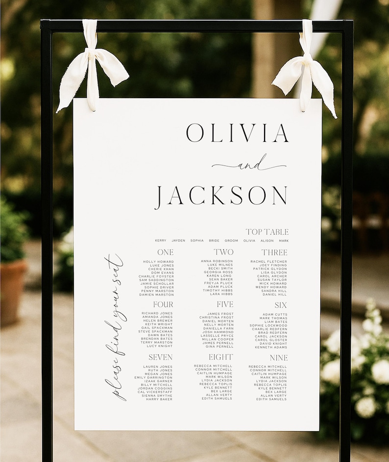 May include: A black metal wedding sign with a white paper insert. The sign features the names "Olivia and Jackson" and a seating chart with guest names. White ribbons adorn the top.