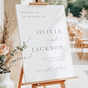 Op de afbeelding: Een wit welkomstbord voor een bruiloft op een houten ezel. De tekst luidt "WELCOME TO THE WEDDING OF OLIVIA and JACKSON", met de locatie en datum eronder. Decoratieve bloemelementen zijn zichtbaar op de achtergrond.