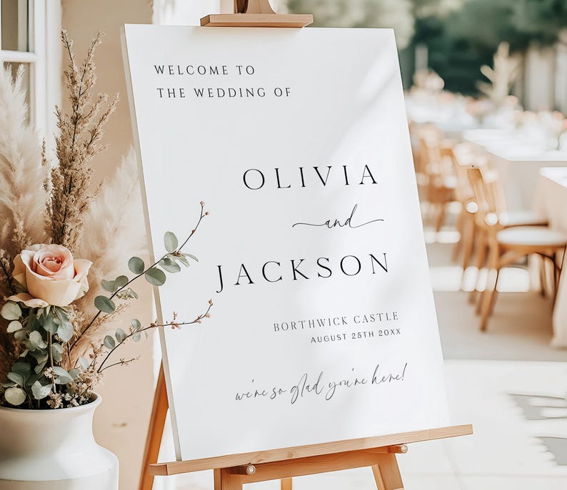 Op de afbeelding: Een wit welkomstbord voor een bruiloft op een houten ezel. De tekst luidt "WELCOME TO THE WEDDING OF OLIVIA and JACKSON", met de locatie en datum eronder. Decoratieve bloemelementen zijn zichtbaar op de achtergrond.
