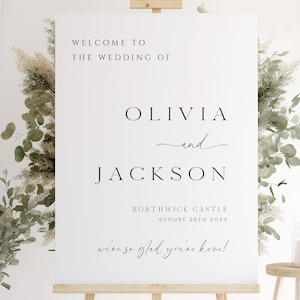 Op de afbeelding: Een witte welkomstbord voor een bruiloft op een houten ezel. Op het bord staat "WELCOME TO THE WEDDING OF OLIVIA and JACKSON" met de locatie en datum eronder. Decoratief groen omringt het bord.