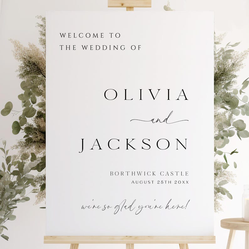 Op de afbeelding: Een witte welkomstbord voor een bruiloft op een houten ezel. Op het bord staat "WELCOME TO THE WEDDING OF OLIVIA and JACKSON" met de locatie en datum eronder. Decoratief groen omringt het bord.