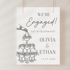 K&ouml;nnte beinhalten: Ein wei&szlig;es Schild mit den Worten "We're Engaged!" in eleganter Schrift, darunter "Let's Celebrate!". Eine Zeichnung zeigt Champagner, der in eine Pyramide aus Gl&auml;sern gegossen wird. Die Namen "Olivia & Ethan" und das Datum "14.07.2026" sind ebenfalls enthalten.
