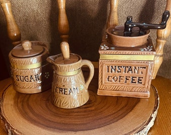 Könnte beinhalten: Ein Set aus drei braunen Keramik-Küchenbehältern mit goldenen Verzierungen und weißen Blumenakzenten. Die Behälter sind mit "Zucker", "Sahne" und "Instantkaffee" beschriftet. Die Behälter stehen auf einem Holztablett.