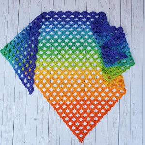 Puede incluir: Un chal de ganchillo arcoíris con un degradado de colores que va del morado al naranja. El chal está hecho con un punto delicado y calado y tiene un borde con flecos.