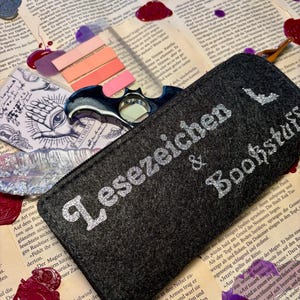 Könnte beinhalten: Eine graue Filz-Stiftetui mit silbernen Glitzerbuchstaben, die "Lesezeichen & Bookstars" lesen.