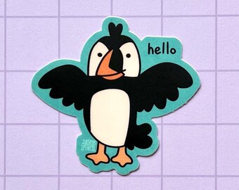 May include: A sticker featuring a cartoon puffin with outstretched wings. The puffin is black, white, and orange, with a teal outline. The word "hello" is written above the puffin. The sticker is on a purple grid background.