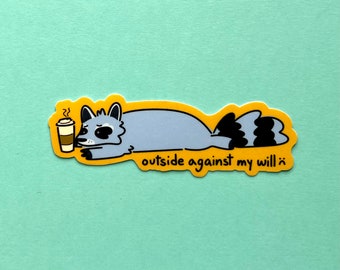 May include: A sticker featuring a cartoon raccoon with a coffee cup, lying down with a tired expression. The text "outside against my will :)" is written in a playful font on a yellow border.
