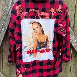 May include: Red and black plaid flannel shirt with a bleached design. The shirt features a graphic of a woman and the text "IN THE STICKS" in red. The shirt is hanging on a black hanger.