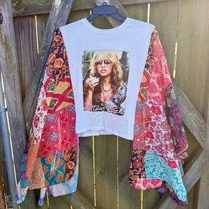 May include: White t-shirt with a graphic of a woman wearing sunglasses, with the text "Stevie Nicks". The sleeves are patchwork with a variety of patterns and colors, including red, pink, blue, and gold.