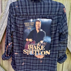 May include: A black and gray plaid flannel shirt with a bleach-splattered design. A square patch with a photo of Blake Shelton and the text "Country Music Freaks Tour 2018" is sewn onto the back of the shirt.