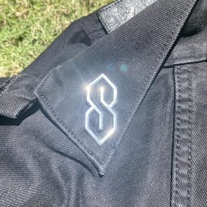 May include: A silver-colored, stylized 'S' pin on a black denim jacket.