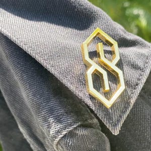 May include: A gold-colored metal pin shaped like the letter 'S' with a geometric design. The pin is attached to a dark gray fabric.
