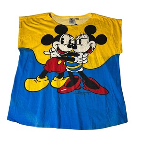 Może przedstawiać: Żółto-niebieska koszulka z grafiką przedstawiającą Myszki Miki i Minnie, które się obejmują. Koszulka ma żółtą górę i niebieski dół, z postaciami z kreskówek pośrodku. Koszulka ma okrągły dekolt.