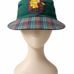 May include: A green baseball cap featuring a plaid brim and a central embroidered patch. The patch displays a yellow cartoon cat character positioned over a large red-outlined letter G, set against a dark green fabric background.
