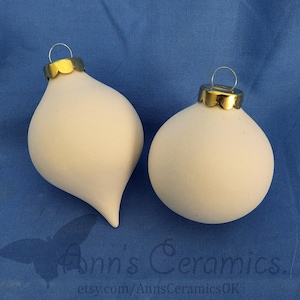 May include: Two white ceramic Christmas ornaments. One is teardrop-shaped, the other is round. Both have gold-colored metal caps with a silver wire loop for hanging. The ornaments are on a blue background. The text "Ann's Ceramics" is visible.