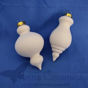 May include: Two white ceramic Christmas ornaments with gold-colored metal caps and wire hangers. One ornament is bulbous, the other is teardrop-shaped with a spiral design. The ornaments are on a blue background.