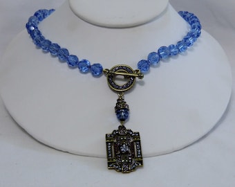 Puede incluir: Un collar de cuentas de cristal azul claro con un colgante dorado en forma de cuadrado con detalles intrincados y un cristal transparente en el centro.