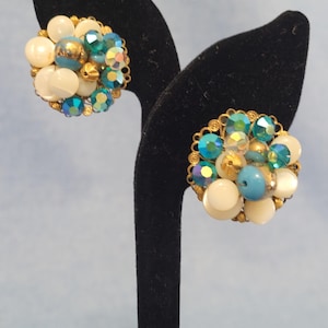 May include: Vintage clip-on earrings featuring a cluster design. The earrings showcase a mix of white, light blue, and iridescent beads, accented with gold-tone metal detailing. The earrings are displayed on a black stand against a light blue background.