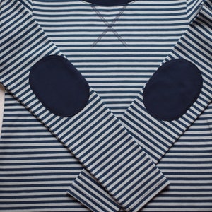 May include: A long-sleeved striped shirt in navy blue and white, featuring navy blue elbow patches. The shirt has a crew neck with a navy blue trim and a stitched detail at the neckline. The brand label is visible.