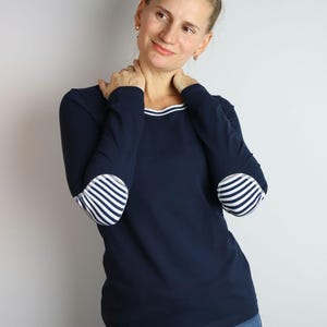 May include: Navy blue long-sleeve shirt with white striped elbow patches and a white neckline trim. The shirt is paired with blue jeans. A woman is wearing the shirt.