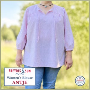 May include: A light lavender blouse with a subtle grid texture, featuring three-quarter length sleeves, a split neckline with thin ties, and a loose fit. The garment is paired with blue denim jeans. Text on the image reads FREYDIS & SUN.