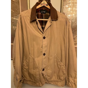 May include: A tan-colored jacket featuring a brown corduroy collar, a button-front closure, and two large front pockets. The interior is lined with a plaid fabric in shades of red, green, and yellow. The label reads J.CREW.