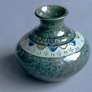 Może przedstawiać: Ręcznie robiony ceramiczny wazon z nakrapianą turkusową glazurą. Wazon posiada ozdobny pas z białymi, niebieskimi i żółtymi geometrycznymi wzorami. Wazon ma zaokrąglony korpus i wąską szyjkę.