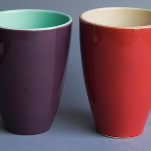 Op de afbeelding: Twee keramische vazen. Eén is dieppaars met een mintgroene binnenkant. De andere is felrood met een crèmekleurige binnenkant. Beide hebben een glanzende afwerking en een eenvoudige, elegante vorm.