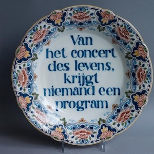Può includere: Un piatto decorativo rotondo in ceramica con bordo smerlato, caratterizzato da un centro bianco con il testo blu "Van het concert des levens, krijgt niemand een program". Il bordo presenta un motivo floreale nei toni del blu, arancione e verde.