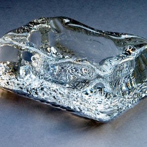 Op de afbeelding: Een heldere, rechthoekige asbak met een getextureerd, geribbeld oppervlak. De asbak heeft een ondiepe, open kom en is gemaakt van een transparant materiaal, waarschijnlijk glas of kristal. Het ontwerp is modern en minimalistisch.