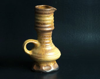 May include: A brown and tan ceramic pitcher with a textured surface and a handle. The pitcher has a wide base and a narrow neck.