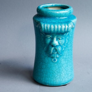 Op de afbeelding: Een turquoise keramische vaas met een gedetailleerd gezichtsontwerp. De vaas heeft een cilindrische vorm met een uitlopende basis en een smalle hals. Het gezicht is gebeeldhouwd met een baard en een hoed. Het oppervlak heeft een craquelé glazuur.