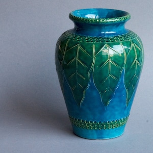 以下が含まれることがあります： 緑の葉のデザインが施されたターコイズ色のセラミック花瓶。花瓶は丸みを帯びたボディと細い首をしています。