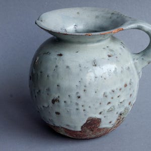 Może przedstawiać: Dzbanek ceramiczny w jasnoniebieskim kolorze z pstrokatą glazurą i brązową krawędzią. Dzbanek ma zaokrąglone ciało i uchwyt.