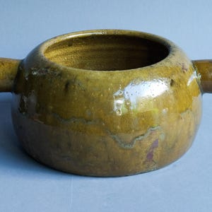 Może przedstawiać: Okrągła ceramiczna miska z dwoma bocznymi uchwytami. Miska ma szeroki otwór i błyszczące, nakrapiane wykończenie w odcieniach oliwkowej zieleni i brązu. Wnętrze ma ciemniejszy, złoty odcień.