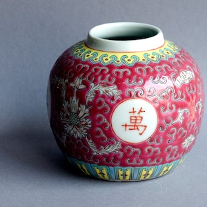 Könnte beinhalten: Eine runde, rote Keramikvase mit einer weiten Öffnung und einem weißen Innenraum. Die Vase hat ein Blumen- und Schriftrollenmuster mit dem chinesischen Schriftzeichen "Wan" in einem weißen Kreis. Der Rand ist gelb und der Boden hat ein blaues und gelbes Muster.