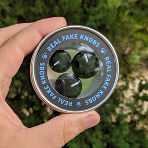 May include: A round metal container with a silver lid and black knobs inside. The lid has the text "REAL FAKE KNOBS" printed on it.