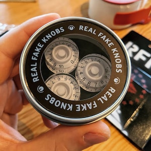 May include: A round tin container with a black lid and white lettering that reads "Real Fake Knobs" on the lid. The container holds three white guitar knobs with black numbers.