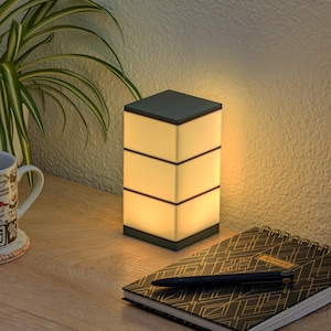 Pode incluir: Uma moderna luminária de mesa com parte superior e base cinza escuro e três seções retangulares brilhantes. Uma caneca com tema de sushi está em uma base, ao lado de um caderno e uma caneta. Uma planta em vaso está ao fundo.