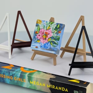 May include: A miniature painting of pink flowers in a blue vase on a small wooden easel. The painting is surrounded by other small easels in white, brown, tan, and black. A book titled "Missing Girls" by Megan Miranda is in the foreground.